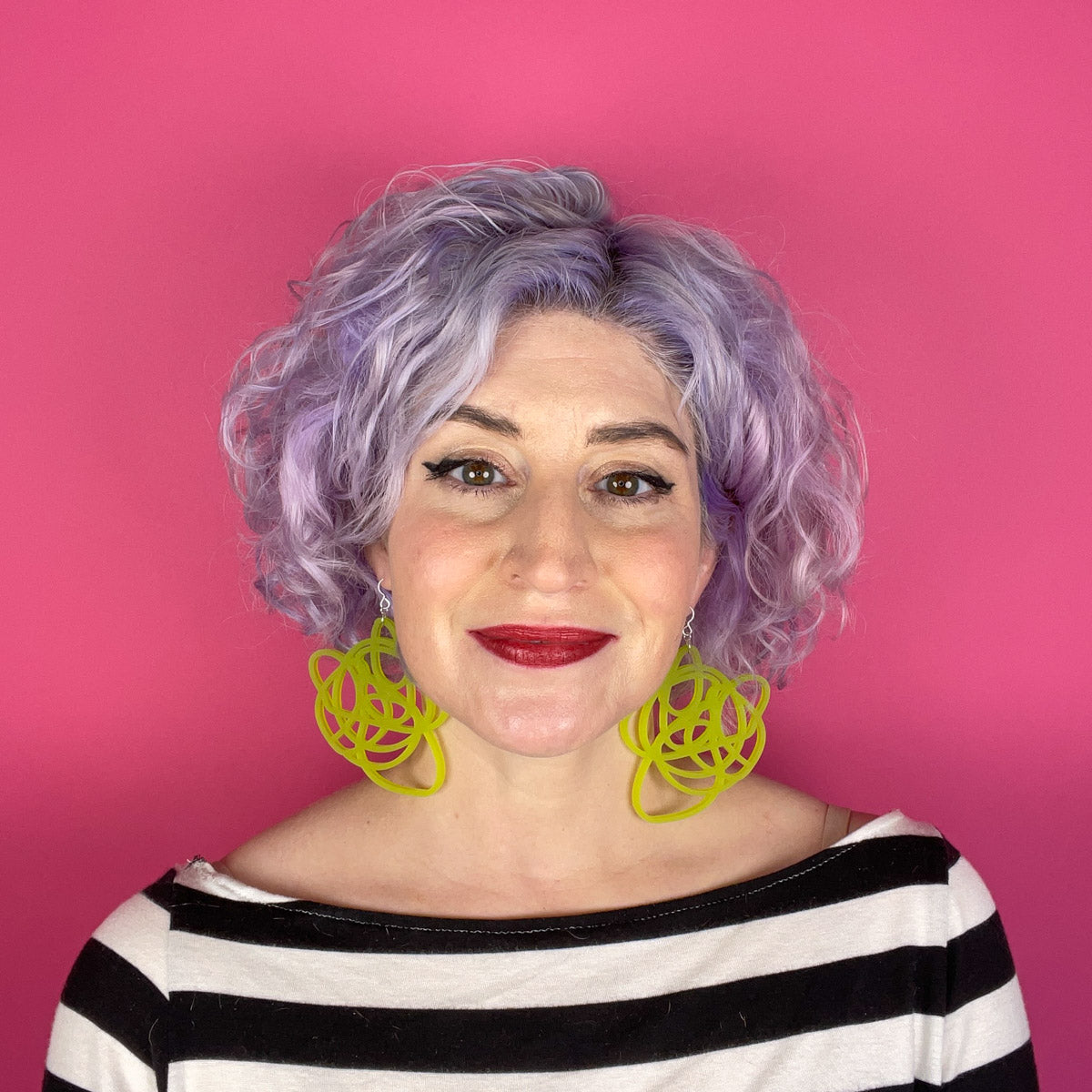 White woman with purple hair wearing lime green statement earrings.