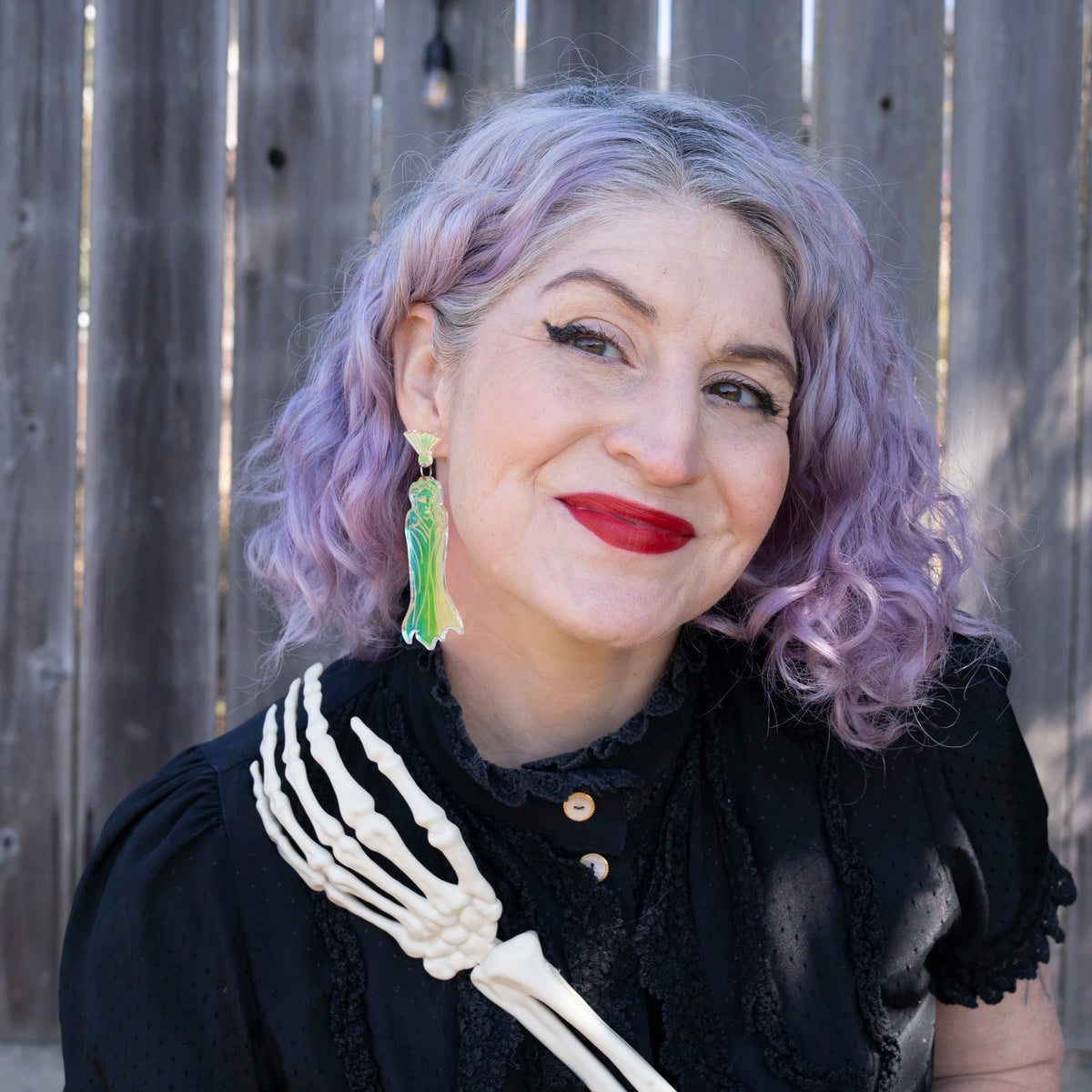 A woman with lavender-purple curly hair models the iridescent ghost earrings wearing a black ruffled blouse and a skeleton hand accessory, standing outdoors in front of a weathered wooden fence.
