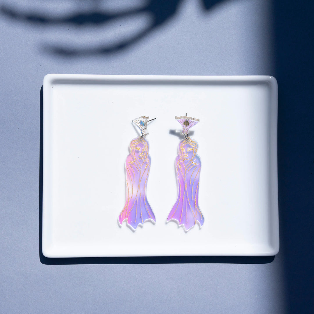 A pair of dangling earrings displayed on a white rectangular tray against a blue-gray background with soft shadows. The earrings have iridescent, holographic pink and purple ghost-like drape shapes with silver-toned metal posts.