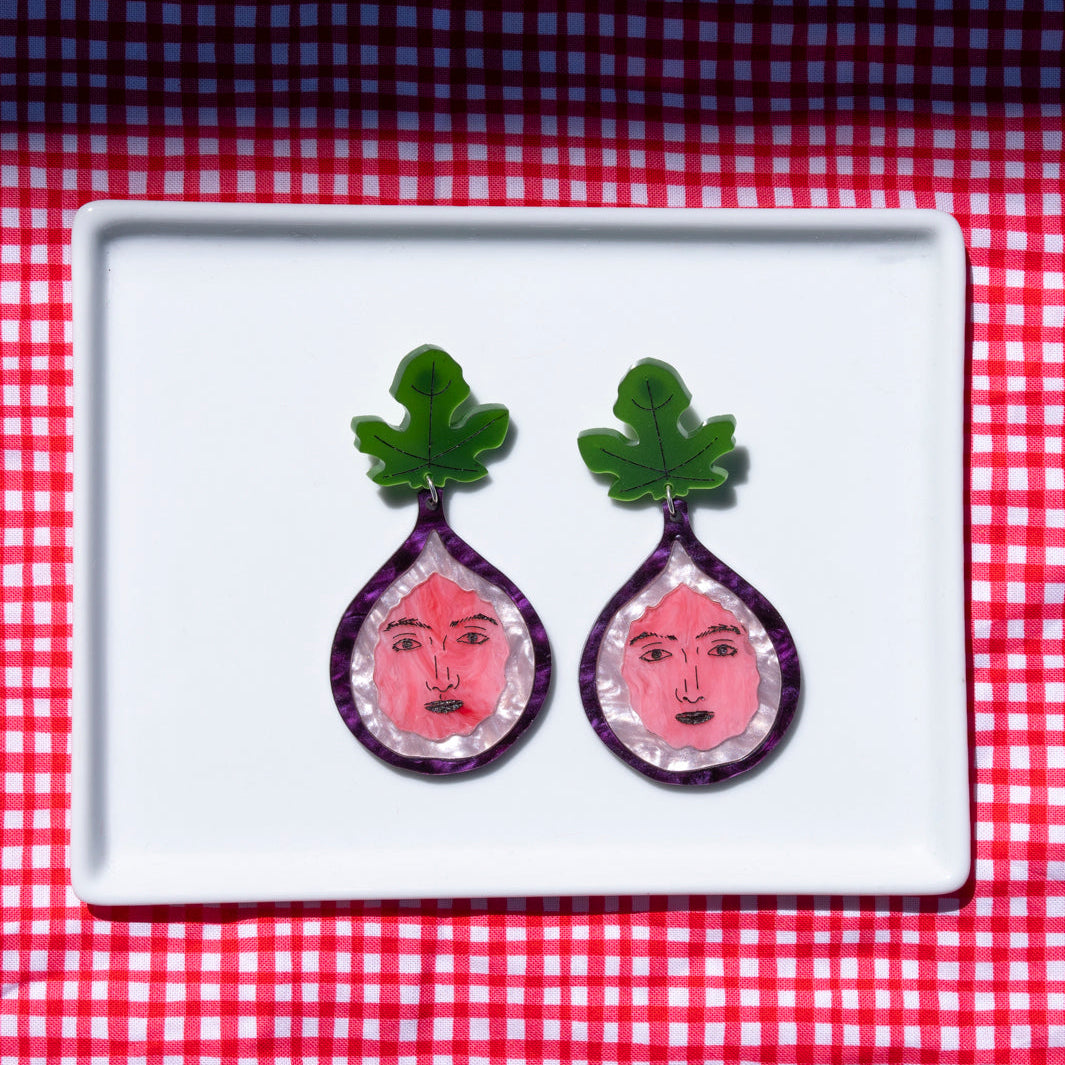 A pair of handcrafted fig face earrings displayed on a white plate against a red and white checkered background.