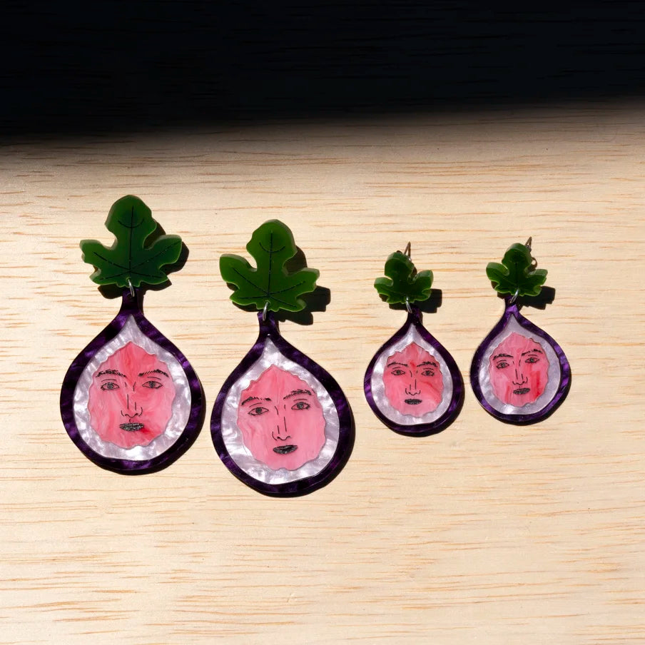 Four fig earrings, each containing a pink face illustration on a light wood background.