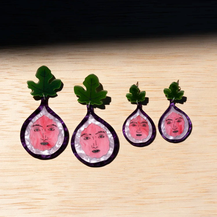 Four fig earrings, each containing a pink face illustration on a light wood background.