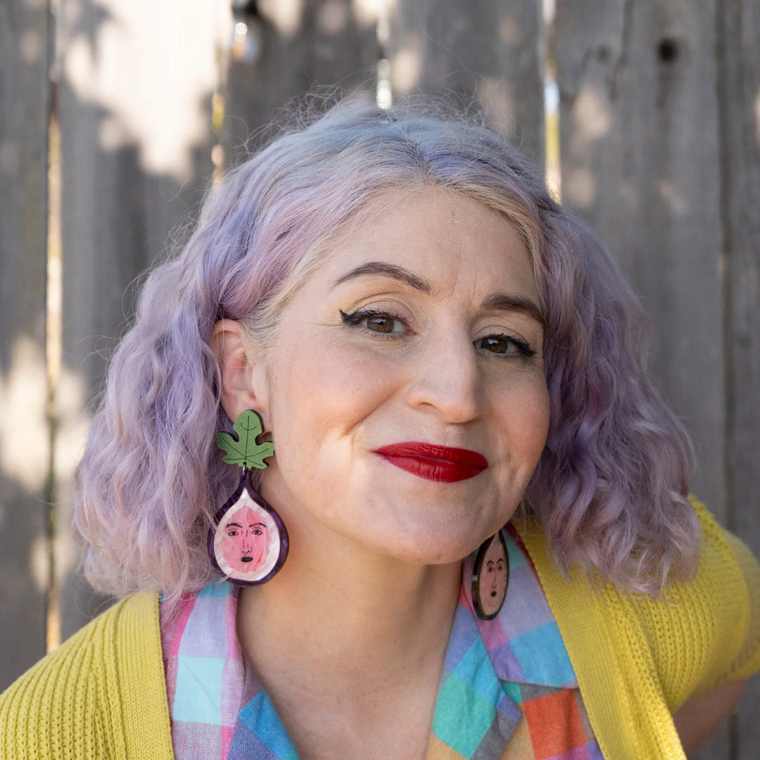 A close-up portrait of a woman with lavender hair wearing handmade fig earrings with faces, a yellow cardigan, and a pastel plaid shirt, standing in front of a weathered wooden fence.