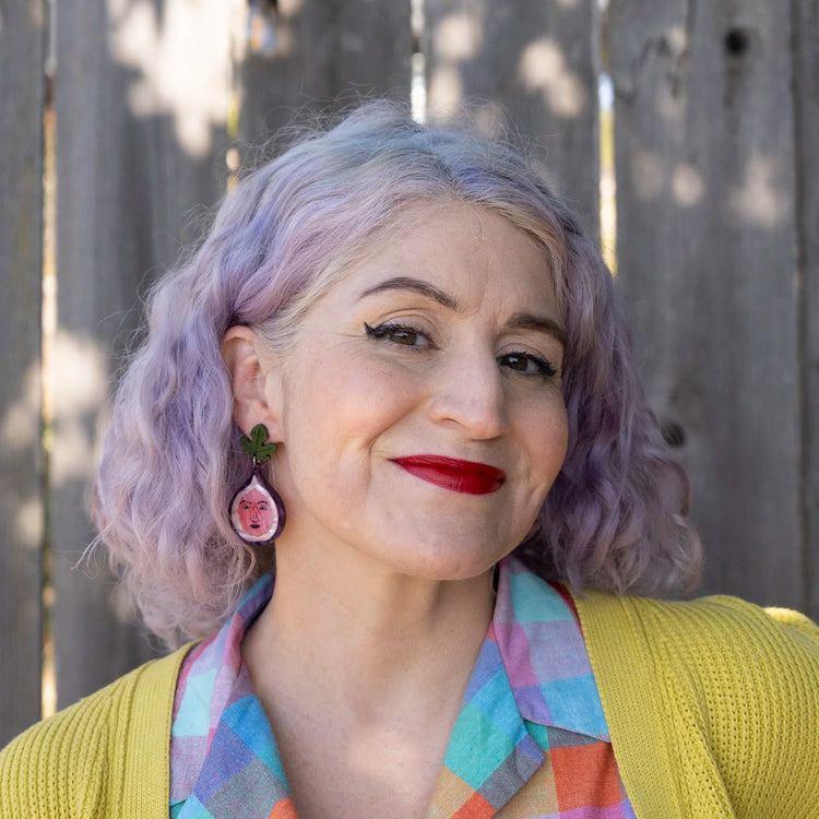 A close-up portrait of a woman with lavender-purple hair wearing a yellow cardigan, a colorful plaid shirt, and handmade fig earring, set against a weathered wooden fence background.