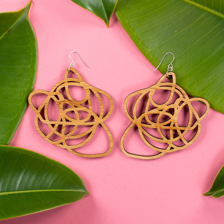 Lightweight wooden graffiti earrings shown on pink background surrounded by leaves. 