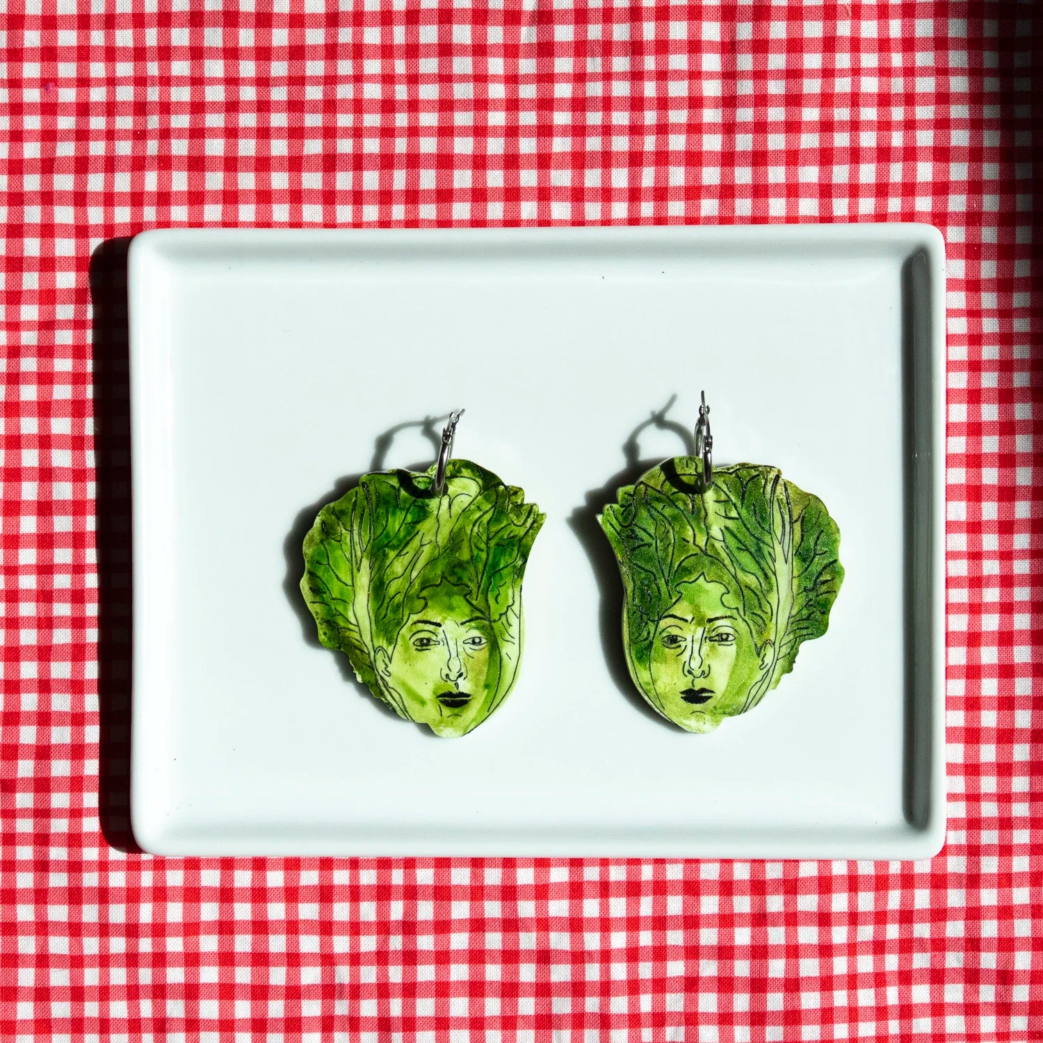 A pair of green lettuce leaf earrings with illustrated faces, displayed on a white rectangular plate over a red and white checkered tablecloth.