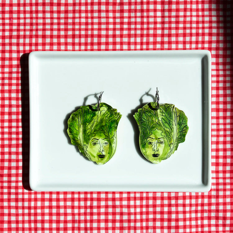 A pair of green lettuce leaf earrings with illustrated faces, displayed on a white rectangular plate over a red and white checkered tablecloth.