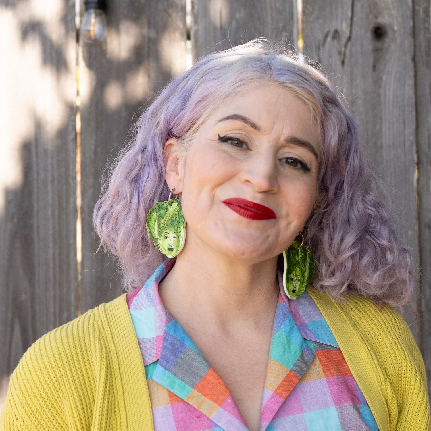A woman with lavender hair and red lipstick wearing large green lettuce leaf-shaped earrings, a yellow cardigan, and a pastel plaid shirt, standing in front of a weathered wooden fence.