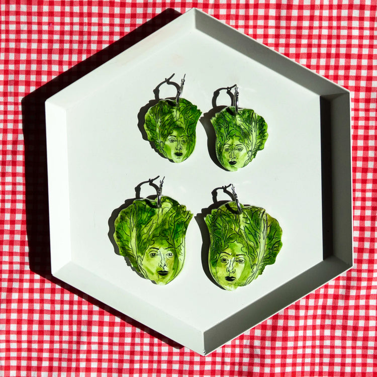 Four green lettuce-shaped earrings with painted faces are arranged on a white hexagonal tray over a red and white checkered tablecloth.