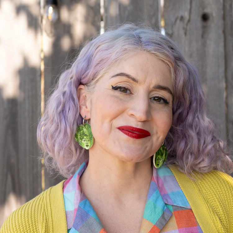 A close-up portrait of a woman with lavender-purple hair wearing large green lettuce leaf-shaped earrings, a yellow cardigan, and a colorful plaid shirt, standing in front of a weathered wooden fence.