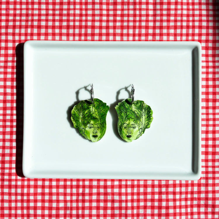 A pair of green lettuce-shaped dangle earrings with painted faces, displayed on a white rectangular plate over a red and white checkered tablecloth.
