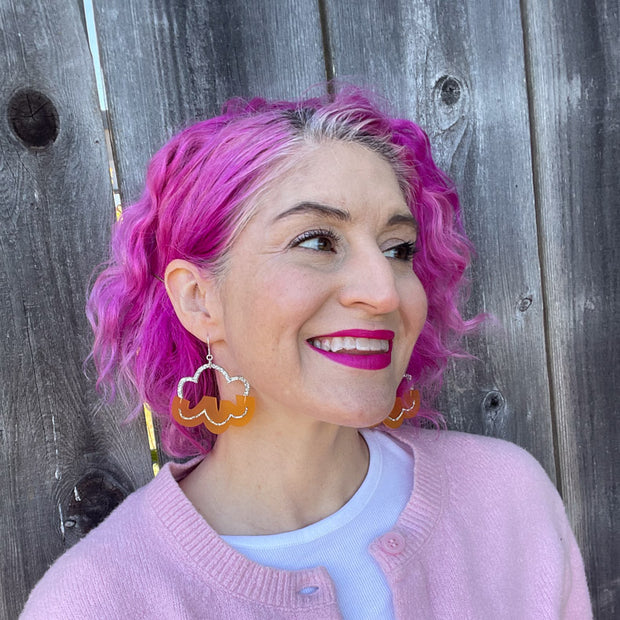 orange ands silver cloud earrings shown on white model with pink hair against a wooden fence