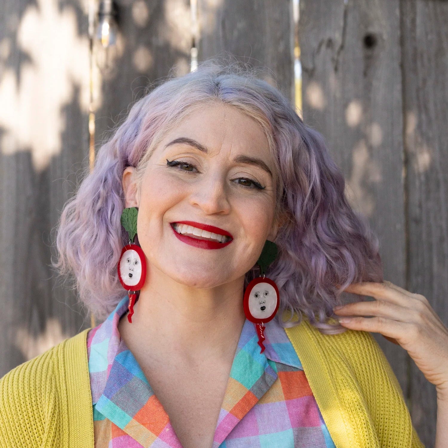 A woman with lavender hair wearing large radish earrings, a colorful plaid shirt, and a yellow cardigan, posing in front of a weathered wooden fence.