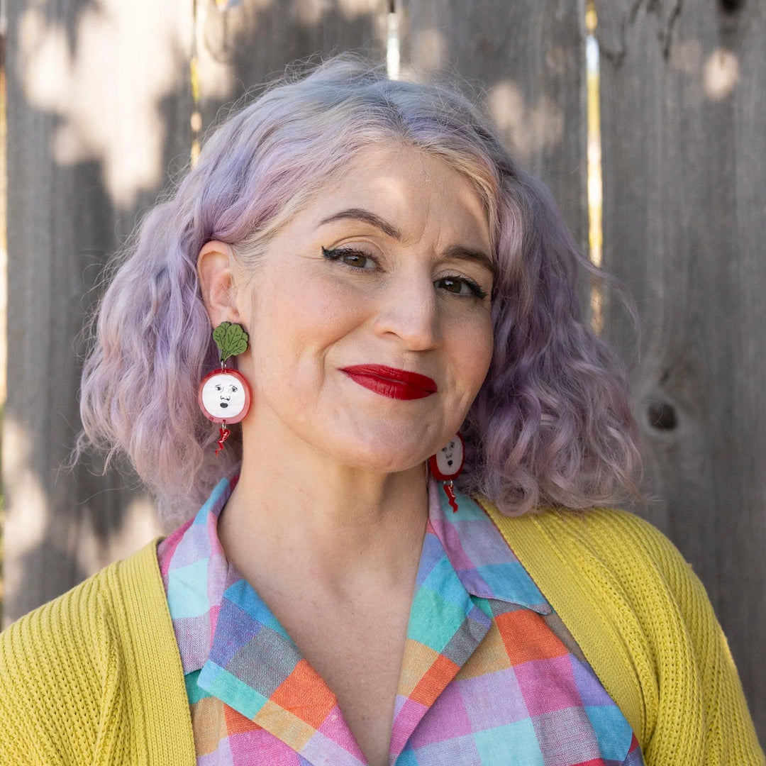 A woman with lavender-purple hair wearing radish earrings, a yellow cardigan, and a pastel plaid shirt, posing in front of a weathered wooden fence.
