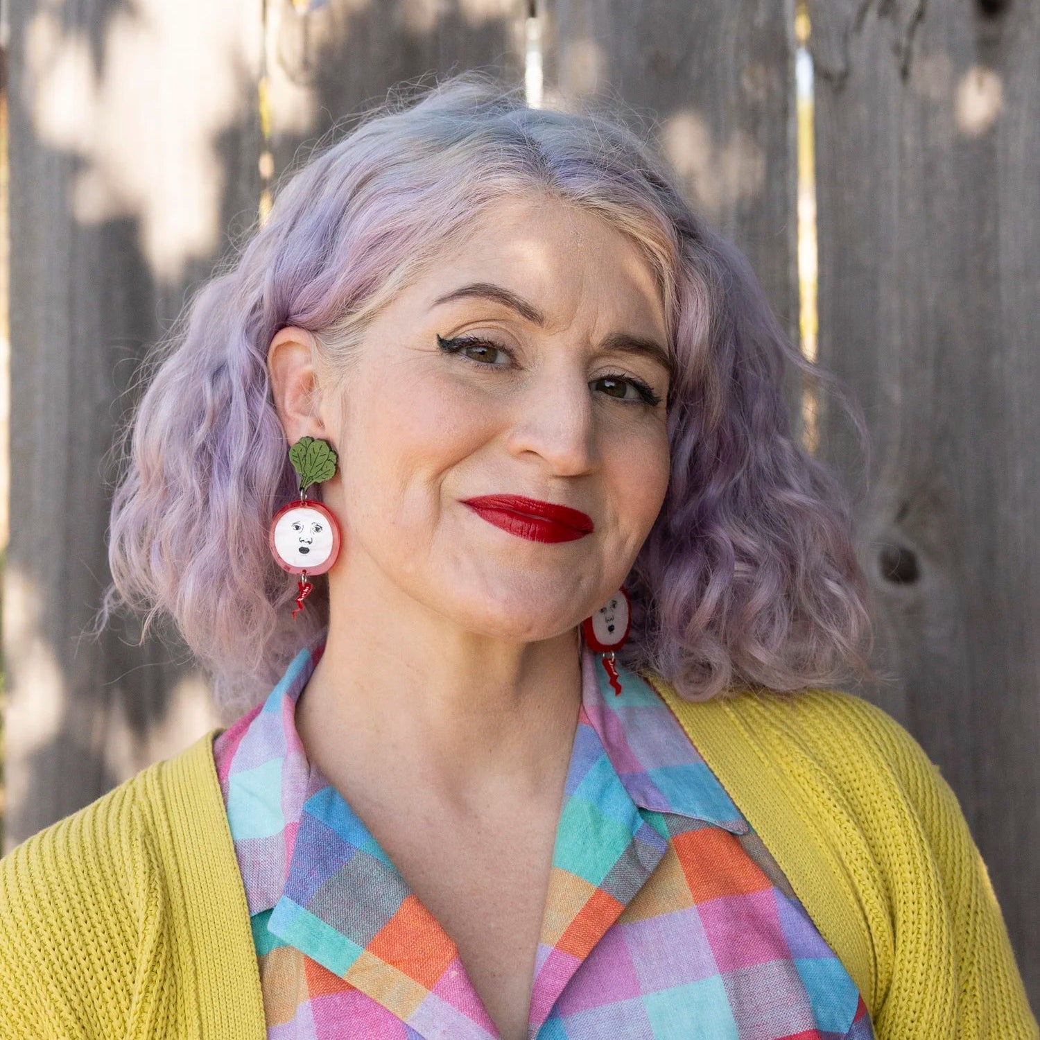 A woman with lavender-purple hair wearing radish earrings, a yellow cardigan, and a pastel plaid shirt, posing in front of a weathered wooden fence.