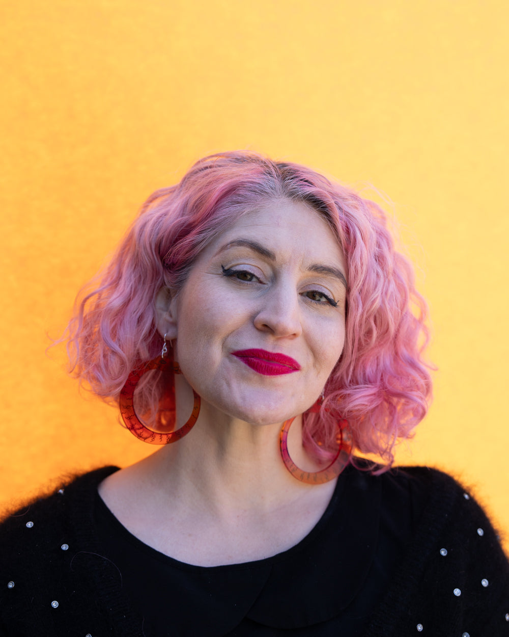 Model with pink hair wearing large red orange ouroboros earrings in front of yellow wall