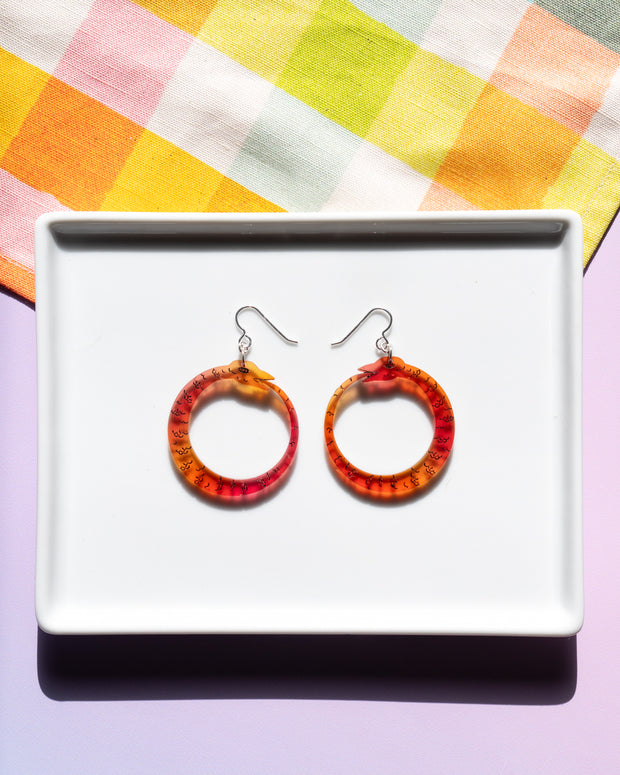 small red orange ouroboros earrings on white tray atop colorful background