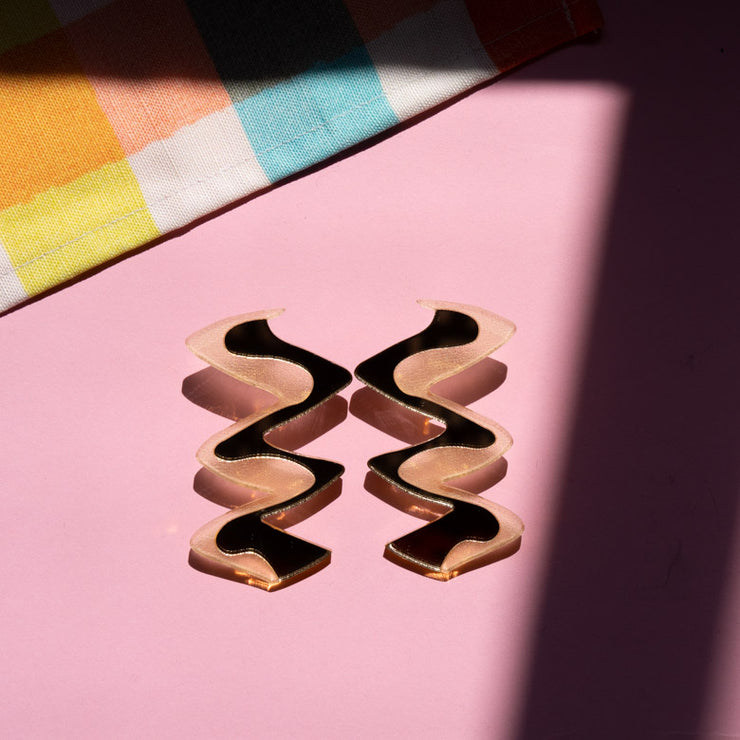 Zigzag gold acrylic earrings shown on pink background
