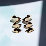 Zigzag gold acrylic earrings shown on light blue background near shadows