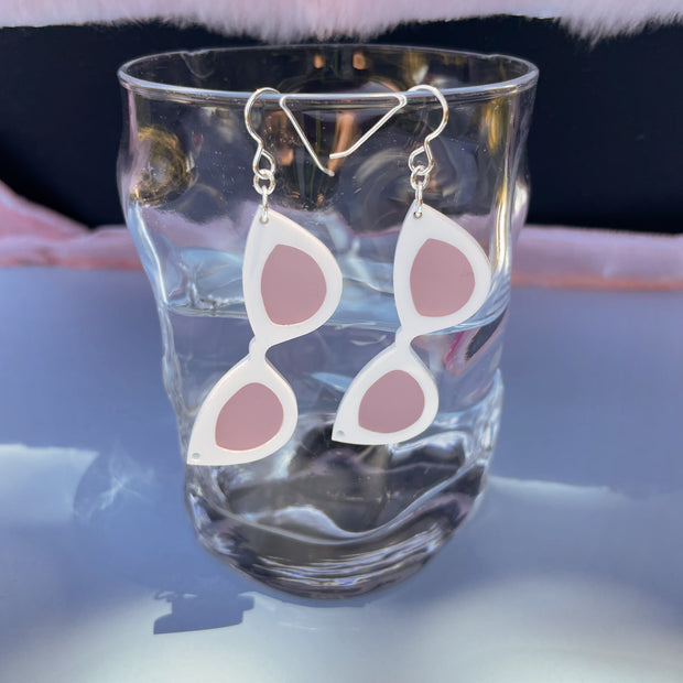 Small white sunglass earrings shown hanging from a glass